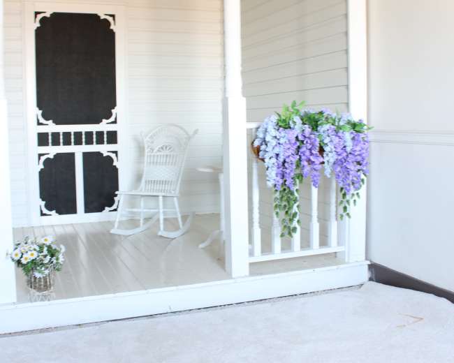 The image shows a porch with a white rocking chair, a decorative black door, and hanging flowers in shades of purple.