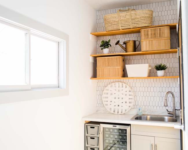 The image shows a minimalist kitchen corner featuring a window, open shelves with woven baskets, a sink, and a small refrigerator beneath the counter.