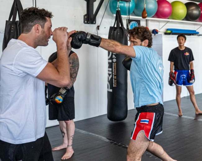 Two athletes practice boxing techniques while two others observe in a training gym.