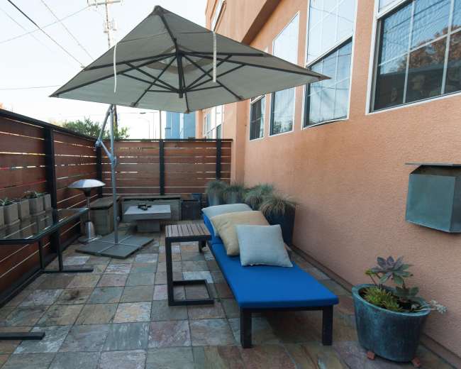 The image shows a small outdoor patio area with a blue bench, potted plants, and a shaded table under an umbrella.