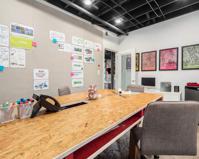 A modern meeting room features a large wooden table surrounded by chairs, with walls displaying colorful project papers and artwork.