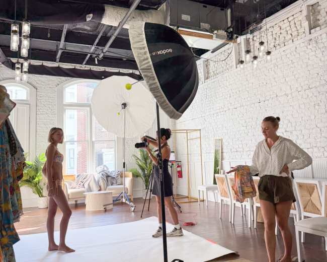 A photography studio features a model posing on a white backdrop while a photographer and an assistant prepare lighting equipment.