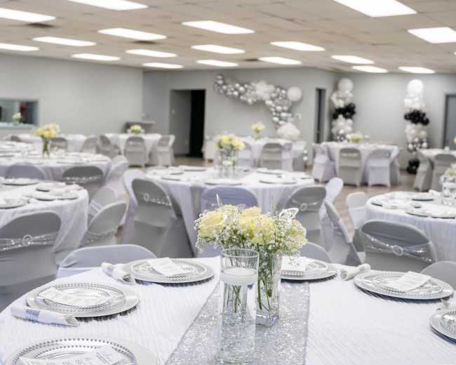 A banquet hall set up for an event with round tables covered in white and silver tablecloths, adorned with floral centerpieces and decorative place settings.