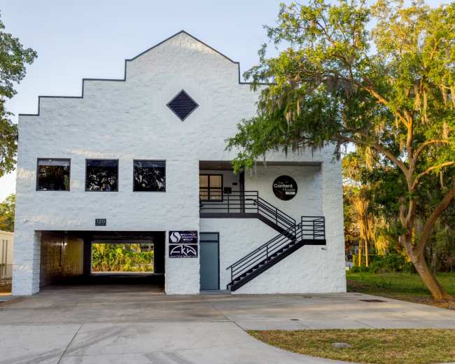 The image shows a modern white building with a distinctive architectural style, featuring a stairway and signage on its facade.