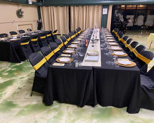 A long banquet table is set with plates and glasses, surrounded by black chairs adorned with yellow sashes, in a spacious room with beige curtains.