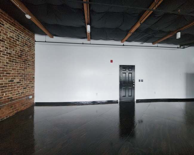 The image shows a spacious room with a black door, exposed brick wall, and dark wooden flooring.