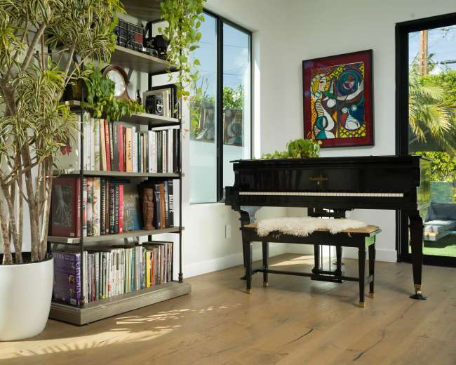 A black grand piano is positioned beside a bookshelf filled with books and surrounded by plants in a well-lit room with large windows.