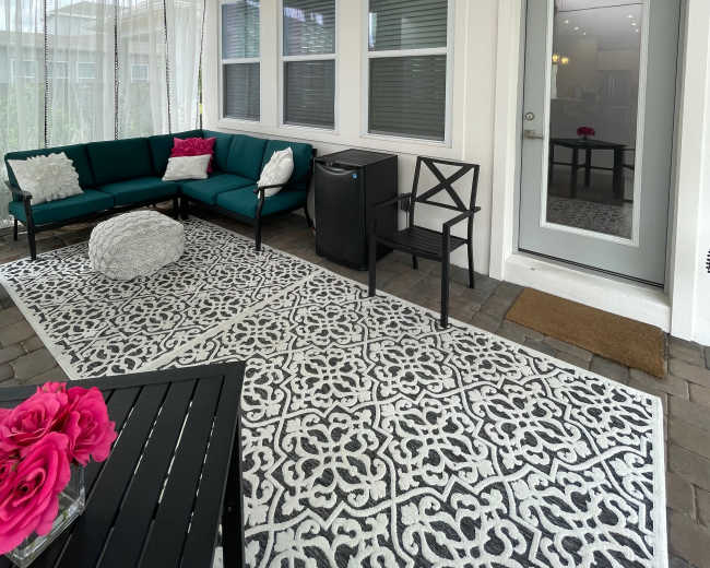 A covered patio area features a green sofa with pink cushions, a round pouf, a small table, and a door leading into an interior space.
