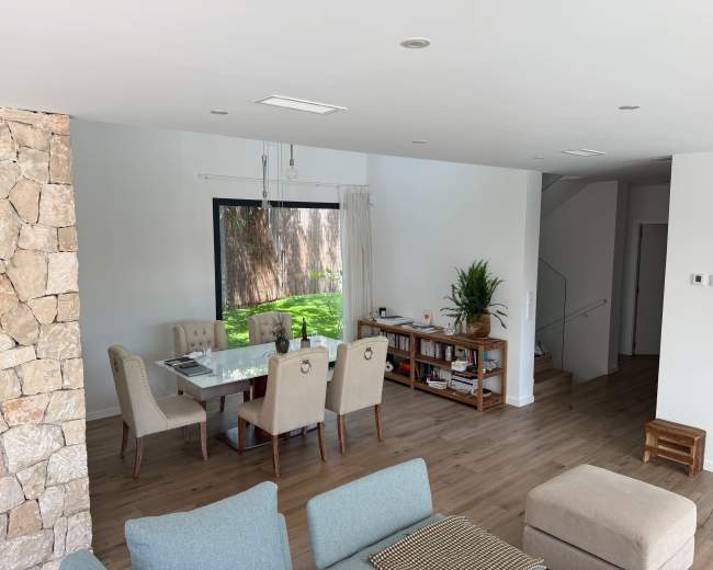 The image shows a modern living room with a dining area featuring a glass table and upholstered chairs, adjacent to a staircase and a large window looking out onto a green space.