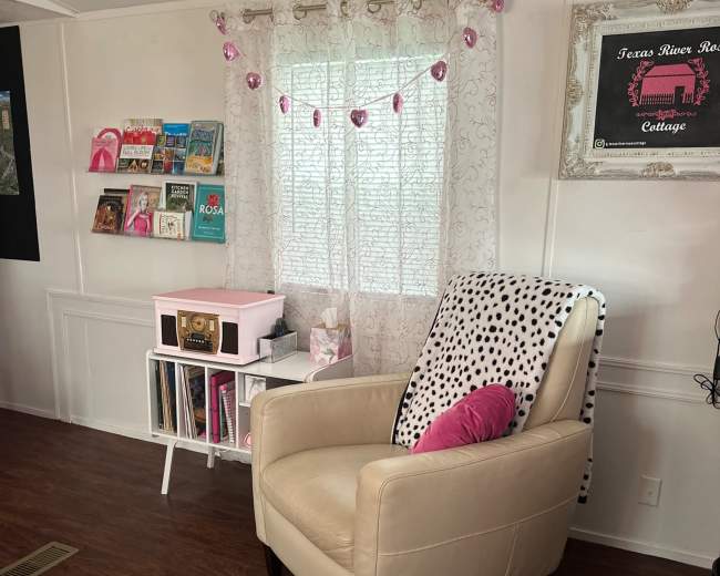 The image shows a cozy reading nook with a beige chair, a pink pillow, a small bookshelf filled with colorful books, and a curtain decorated with pink lights.