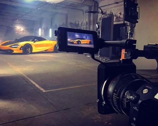 A camera is positioned in front of a yellow McLaren 720S sports car being filmed in a dimly lit industrial setting.