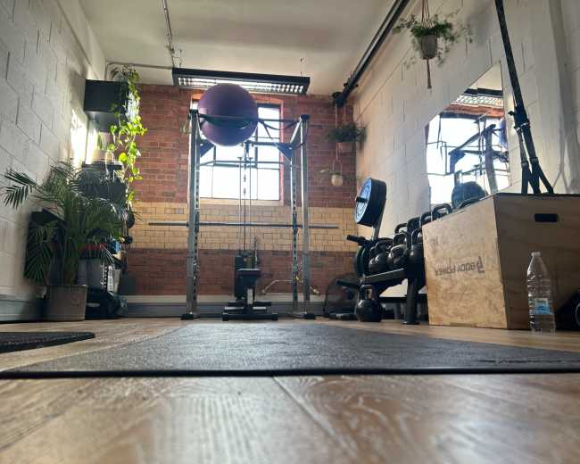 The image shows a gym interior featuring exercise equipment, a strength training rack, a large exercise ball, and a window with natural light illuminating the space.