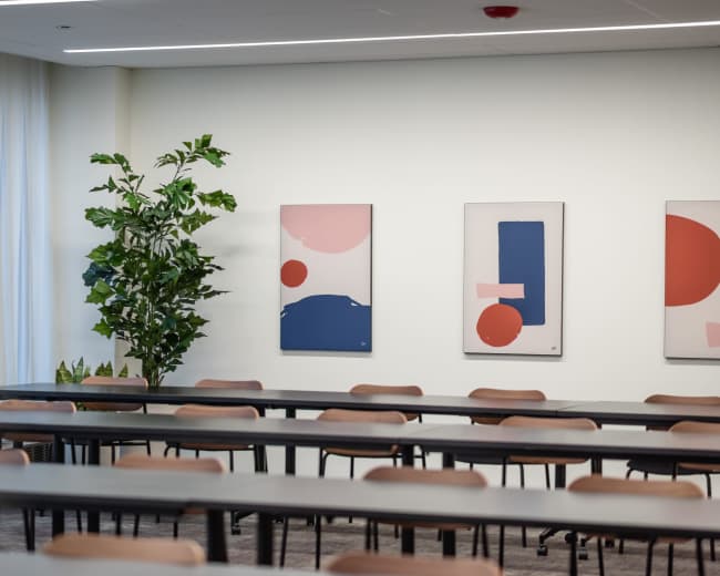 A conference room features rows of tables and chairs, along with abstract wall art and a potted plant in the corner.