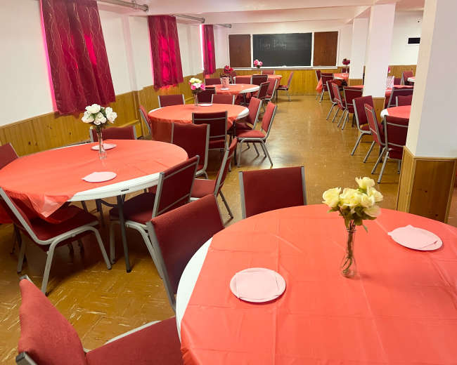 The image shows a banquet room decorated with red tablecloths and centerpieces of flowers on round tables, arranged for an event.