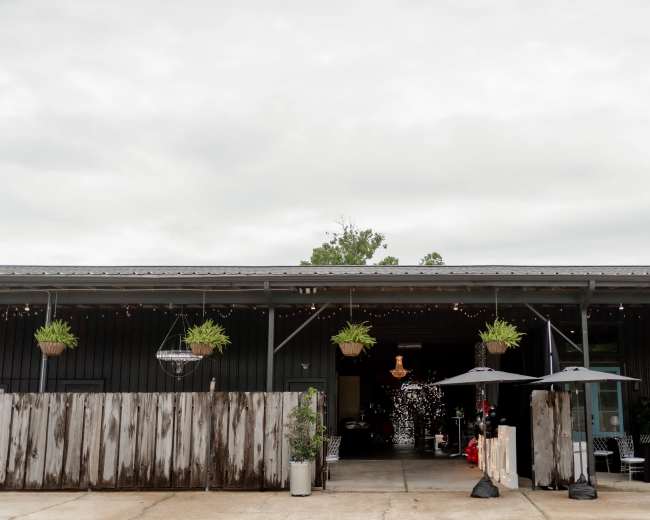 The image shows a rustic building with a dark exterior, featuring hanging plants, outdoor seating with umbrellas, and festive lighting above an entrance.