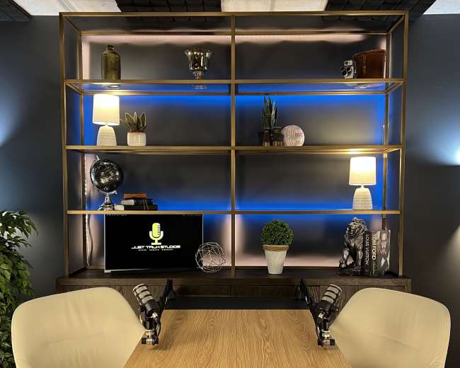 A modern podcast studio features a wooden table with two chairs, flanked by a bookshelf displaying various decorative items, plants, and a computer monitor.