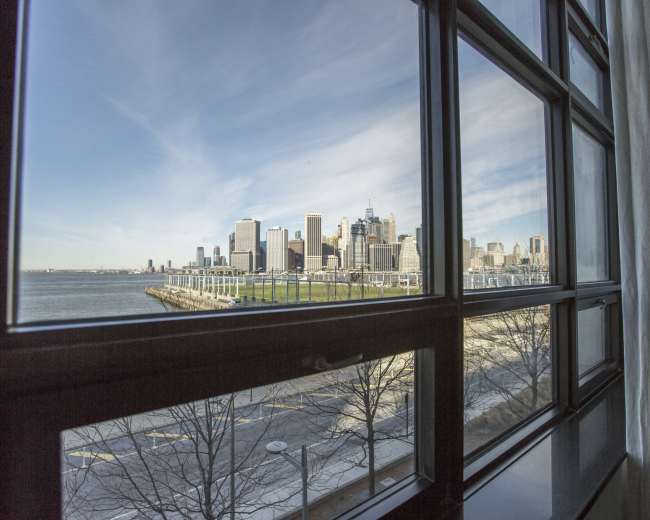 The image shows a view of a city skyline across the water, framed by a window with a partially open curtain.
