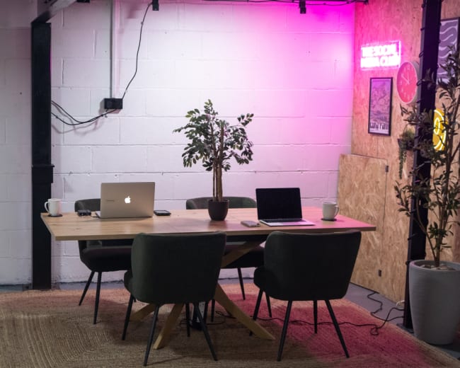A modern meeting room features a wooden table surrounded by four green chairs, with two laptops open on the table and pink ambient lighting in the background.
