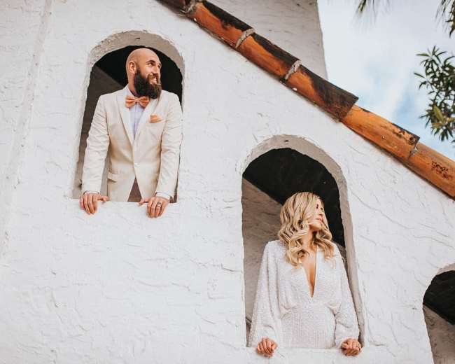 A man in a suit and a woman in a white dress stand in a building with arched openings.