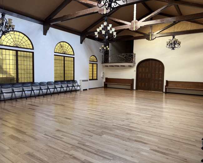 The image shows an empty hall with wooden floors, several folding chairs arranged along the walls, and large arched windows allowing light to enter.