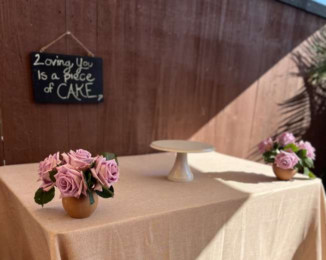 A table with a beige tablecloth is set outdoors, adorned with pink roses in vases and a cake stand, alongside a chalkboard sign that reads "Loving You Is a Piece of Care."