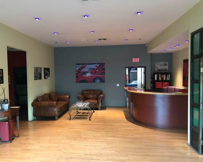 The image shows a reception area with a brown leather sofa, a wooden coffee table, and a curved reception desk, featuring a wall-mounted artwork of cars.