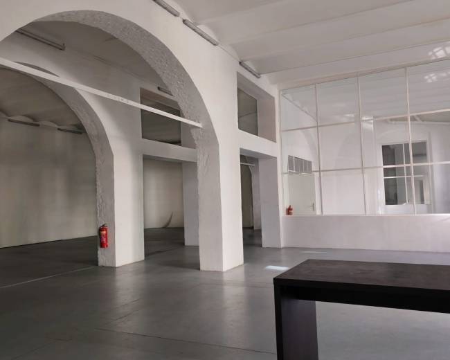 The image shows a spacious interior with white walls, high ceilings, and large arched openings, featuring a few fire extinguishers and minimal furniture.