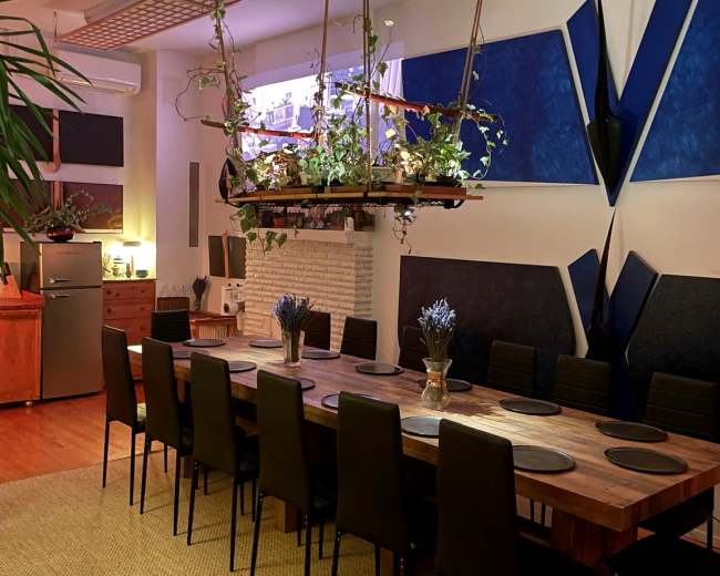 A long dining table is set in a modern room, featuring hanging planters and artistic blue wall decor.