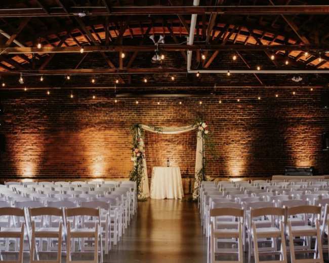 A wedding ceremony setup features rows of white chairs facing a decorated altar against a brick wall in a softly lit venue.