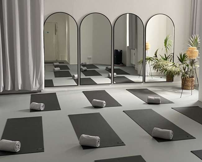 The image shows a minimalist yoga studio arranged with several yoga mats and rolled towels placed in front of three arched mirrors.