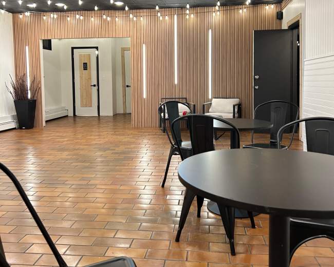 The image shows a spacious interior with wooden paneling, contemporary lighting, and metal chairs arranged around a black table.