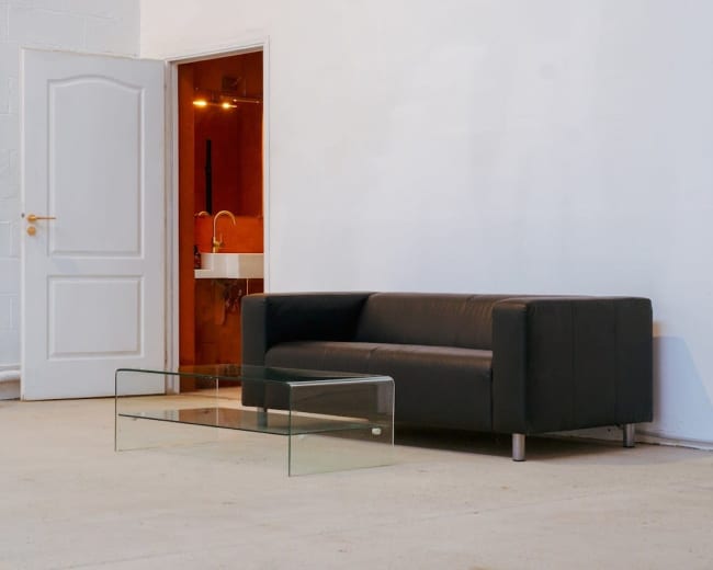A black leather sofa and a glass coffee table are positioned in a minimalist room with an open door leading to a bathroom area.