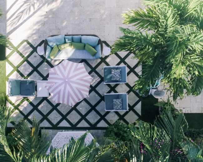 An overhead view shows a patio area featuring a large striped umbrella, four chairs arranged on a geometric patterned rug, and lush greenery surrounding the space.