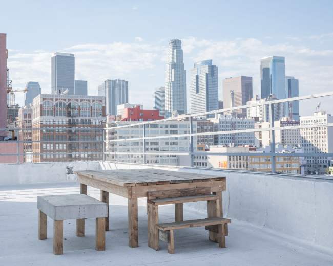 A wooden table and bench are situated on a rooftop overlooking the skyline of downtown Los Angeles.