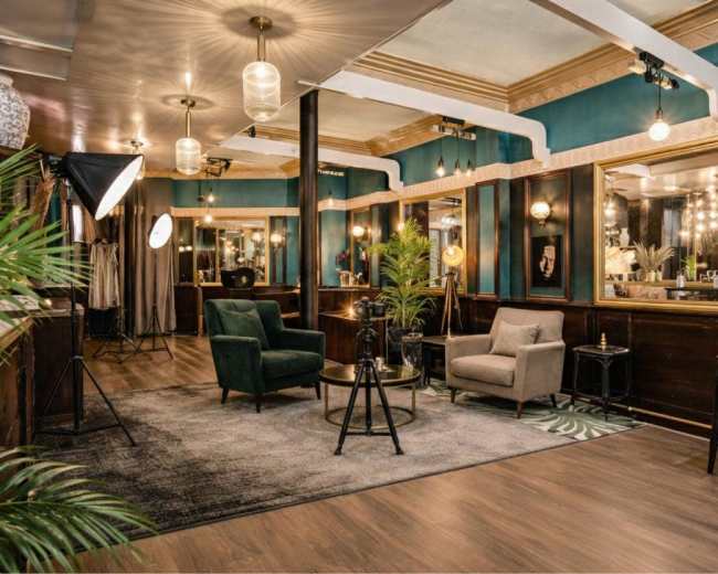 The image shows an elegantly designed interior of a salon featuring two armchairs, a coffee table, and large mirrors, with warm lighting and decorative plants.