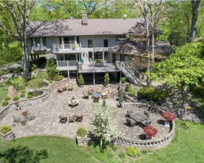 The image shows a spacious home with a multi-level deck overlooking a landscaped backyard featuring a brick patio, seating areas, and a fountain.