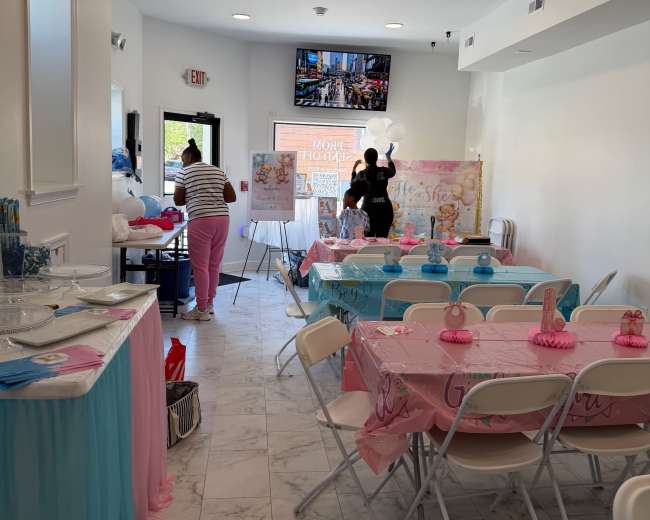 The image shows a decorated event space with tables covered in colorful tablecloths and a large banner at the front, indicating preparations for a celebration.