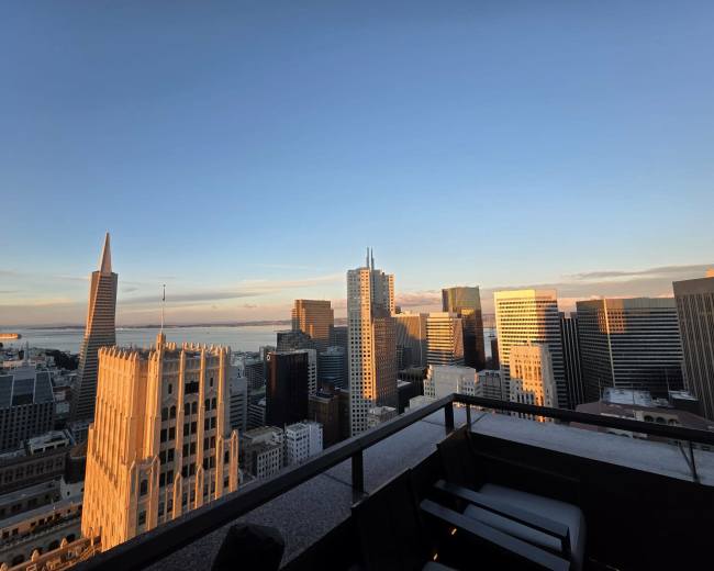 A view from a rooftop terrace overlooking a city skyline at sunset, featuring tall buildings and a glimpse of water in the background.