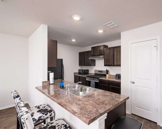 The image shows a modern kitchen with dark cabinetry, a stainless steel stove, and bar seating with patterned stools.