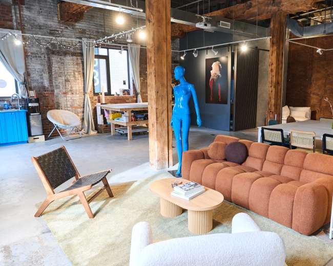 The image shows a modern open-concept living space featuring eclectic furniture, wooden beams, and a blue mannequin.