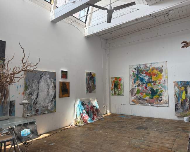 The image shows an artist's studio with colorful abstract paintings hanging on the walls and scattered art materials on the wooden floor.