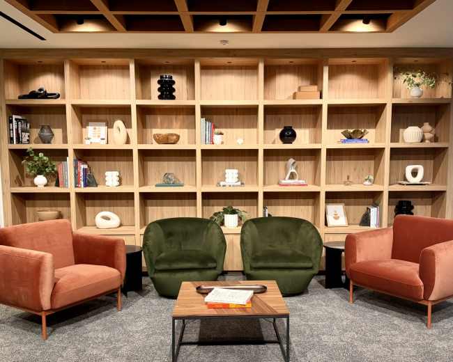 The image shows a cozy lounge area with two upholstered chairs in green and peach, a small wooden coffee table, and a wooden shelving unit filled with various decorative objects and books.