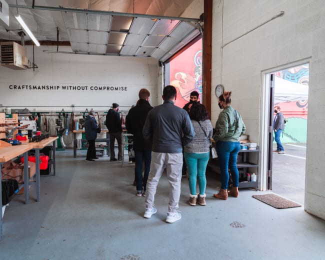 A group of people stands in a workshop, while others browse tools and materials in a well-lit, open space.