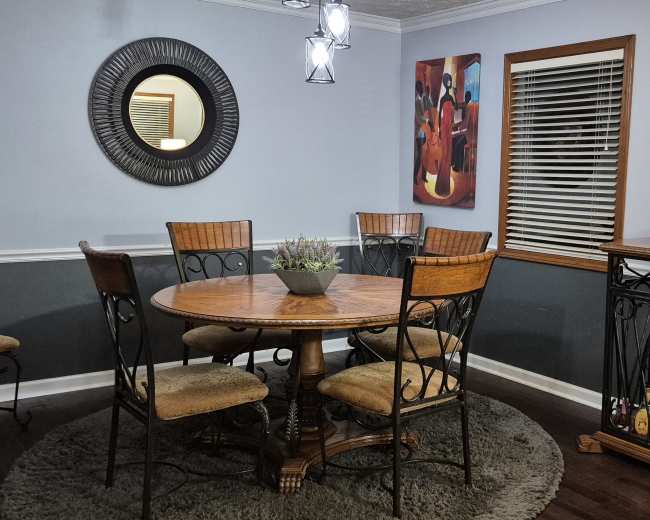 The image shows a dining area with a round wooden table surrounded by six chairs, artwork on the walls, and a bar cabinet in the corner.