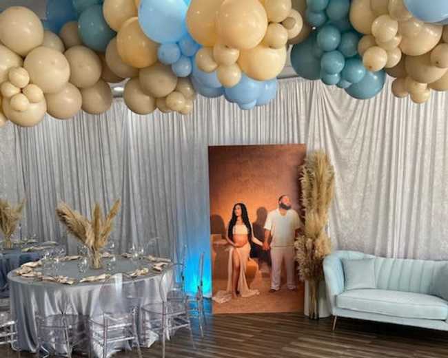 The image shows a decorated indoor event space featuring a balloon arch, a table set for dining, and a large photo backdrop of a couple.