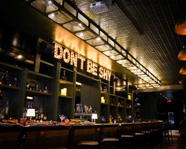 A dimly lit bar interior features a long counter with various bottles displayed on shelves and a neon sign that reads "DON'T BE SHY."
