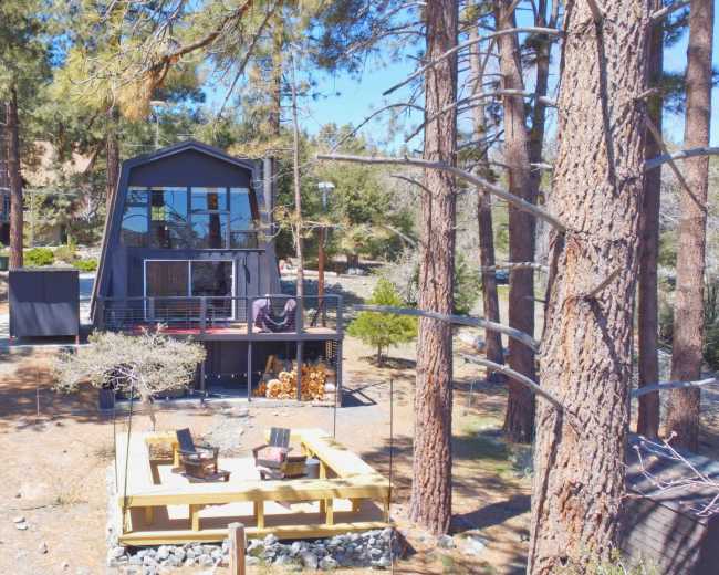 A modern two-story black house with a deck is surrounded by tall pine trees and features a patio area with seating in the foreground.