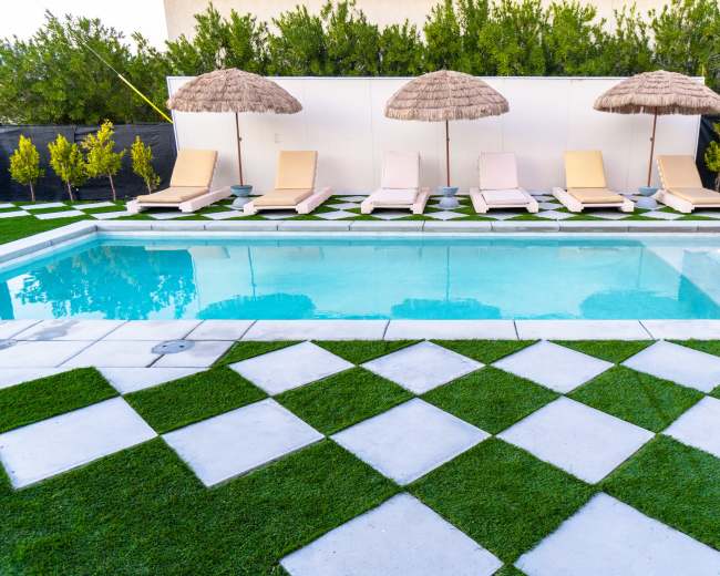 A swimming pool surrounded by lounge chairs and umbrellas, with a neatly arranged grassy area featuring checkered stone tiles.