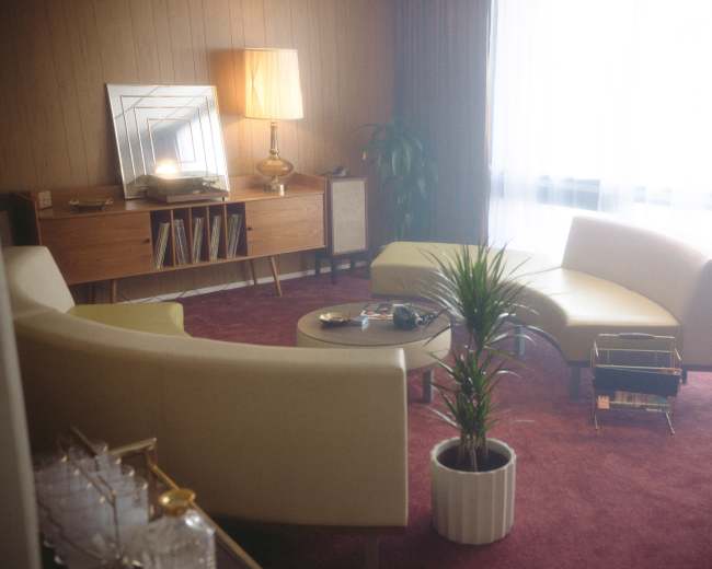 The image shows a mid-century modern living room with curved sofas, a round coffee table, and a decorative lamp, all situated on a carpeted floor.