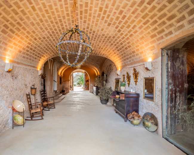 The image shows a spacious, arched hallway with textured stone walls, wooden doors, and decorative elements, leading to an outdoor view.